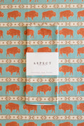 A folded waffle-weave kitchen tea towel with a turquoise blue background, terracotta orange bison print, and cream-and-beige geometric stripes, wrapped with a white paper belly band label that reads 'ASPECT HOME GOODS', laid on a background of the same pattern.
