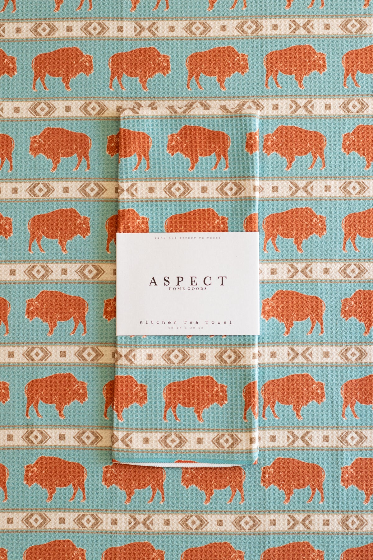 A folded waffle-weave kitchen tea towel with a turquoise blue background, terracotta orange bison print, and cream-and-beige geometric stripes, wrapped with a white paper belly band label that reads 'ASPECT HOME GOODS', laid on a background of the same pattern.
