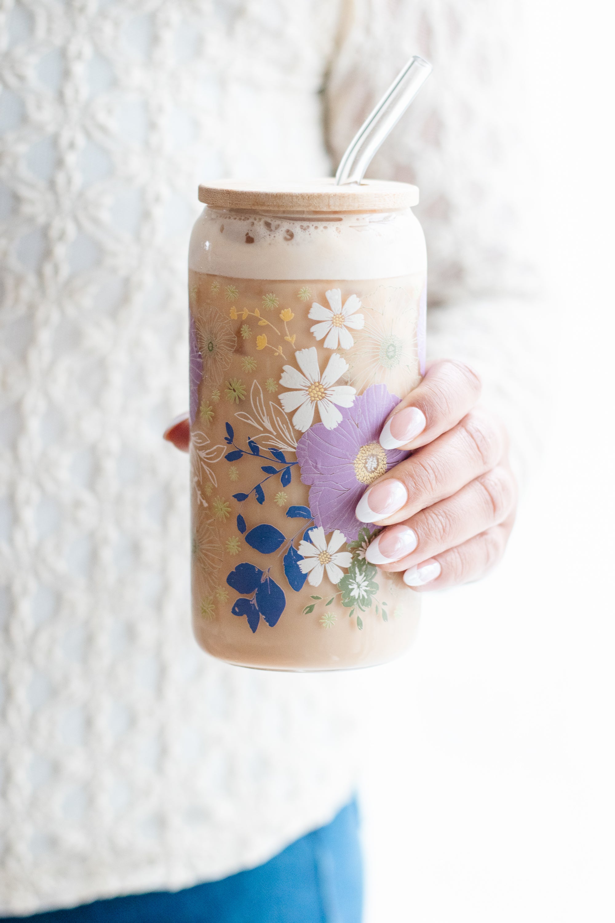 Hand holding a floral-patterned can glass with a glass straw and bamboo lid, against a white background.