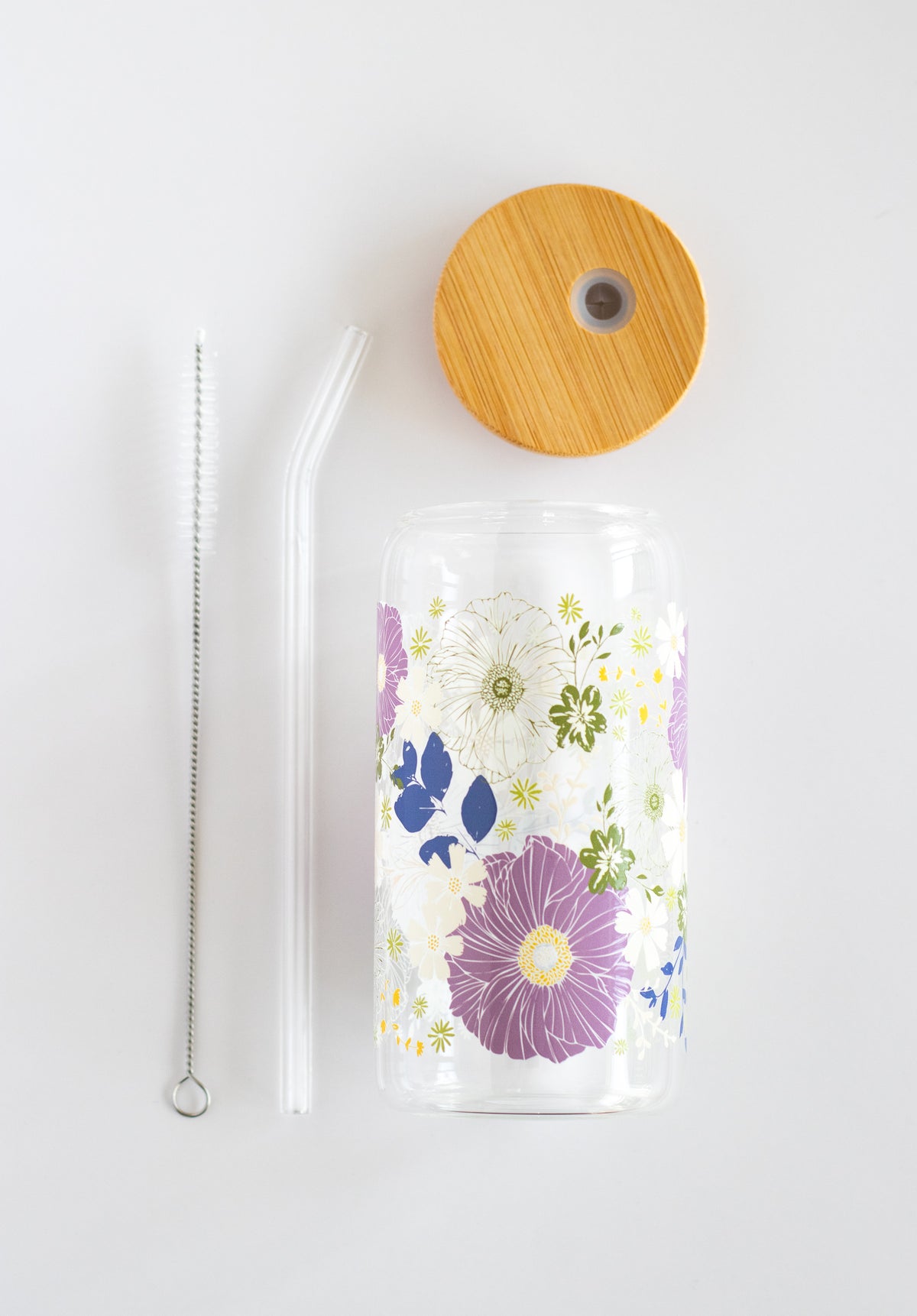 Clear can glass tumbler with a floral design, wooden lid, a glass straw, and a straw cleaning brush, on a white background.