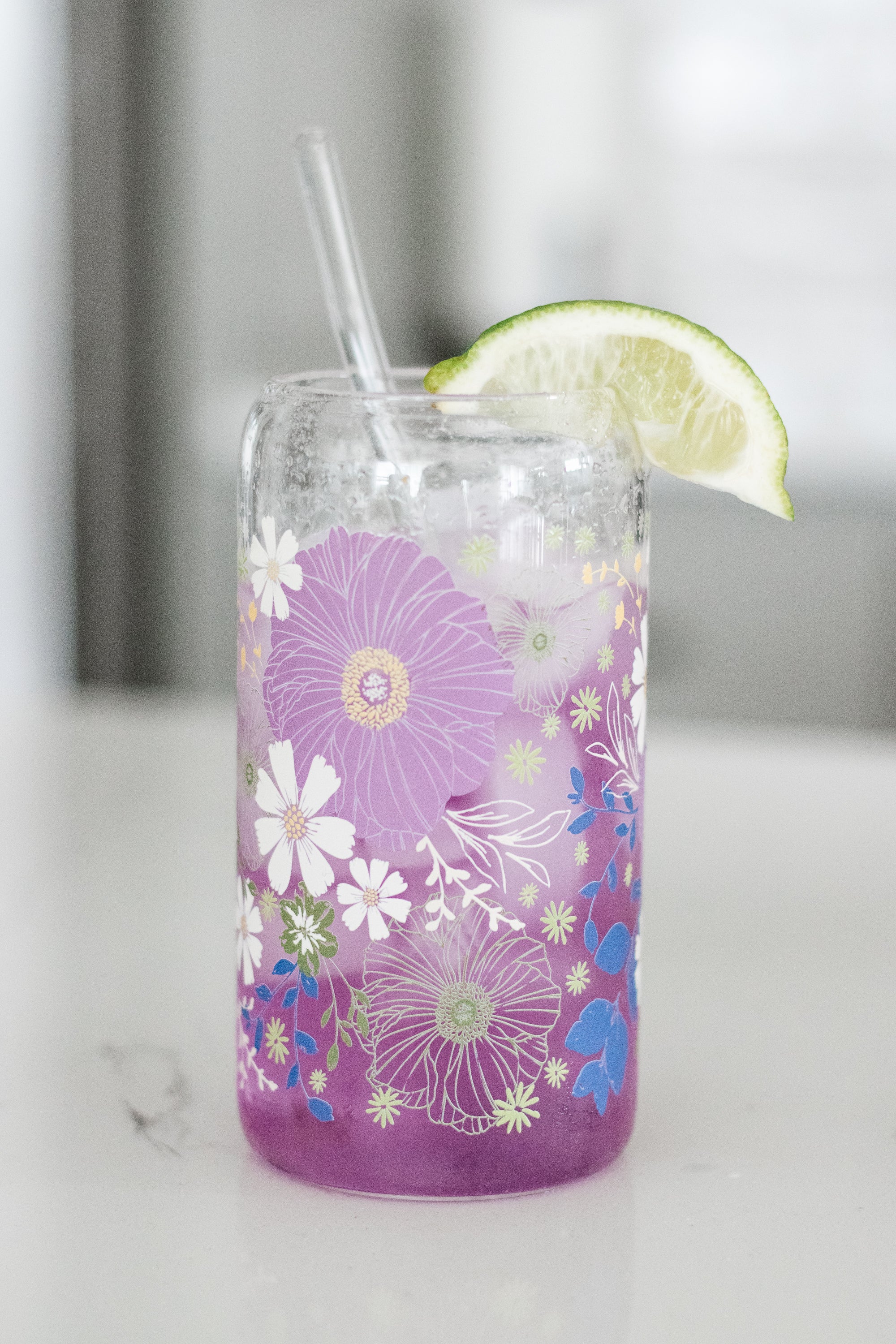 Floral-patterned can glass with a glass straw and a lime wedge, filled with a purple drink, on a white marble surface.