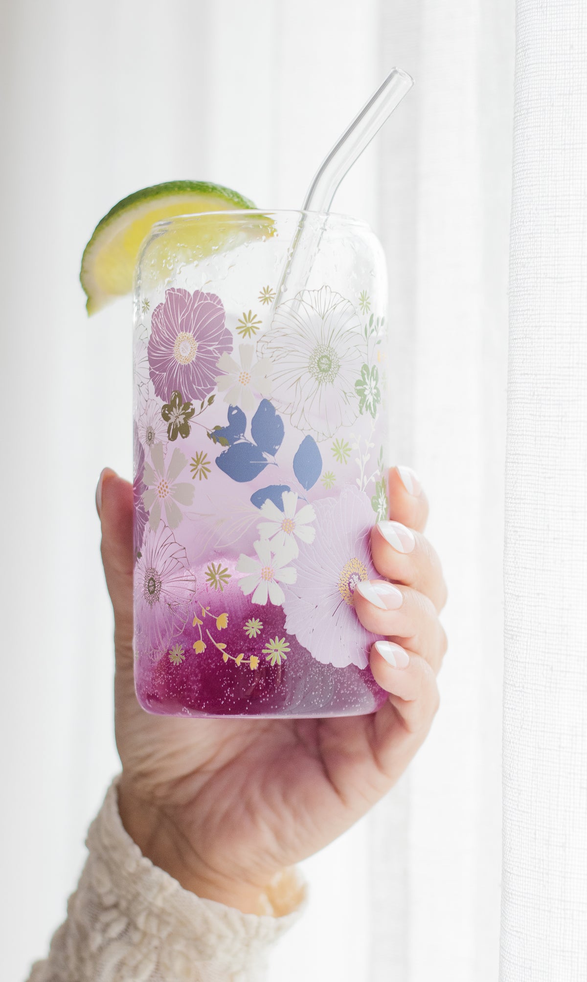Hand holding a floral-patterned can glass with a glass straw and a lime wedge, filled with a purple beverage, against a white background.