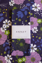 A folded kitchen towel with a floral print lies on top of a matching flat-laid kitchen towel. The pattern features purple, white, and green flowers on a dark navy background, with a white paper belly band around the folded towel reading 'ASPECT HOME GOODS' and 'KITCHEN TEA TOWEL'.