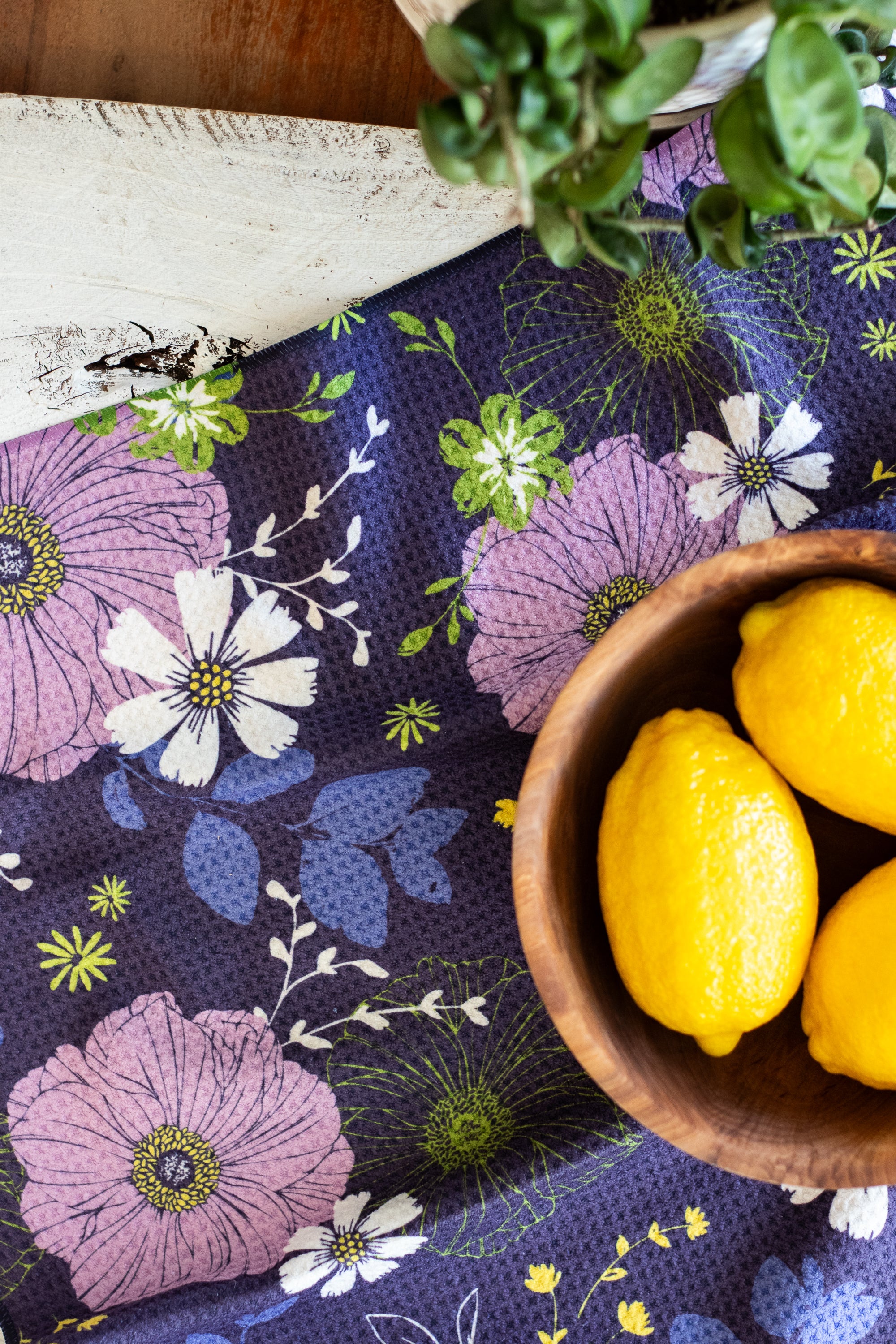 Wooden bowl with lemons and a plant on a floral-patterned waffle-weave kitchen tea towel, against a wooden surface.