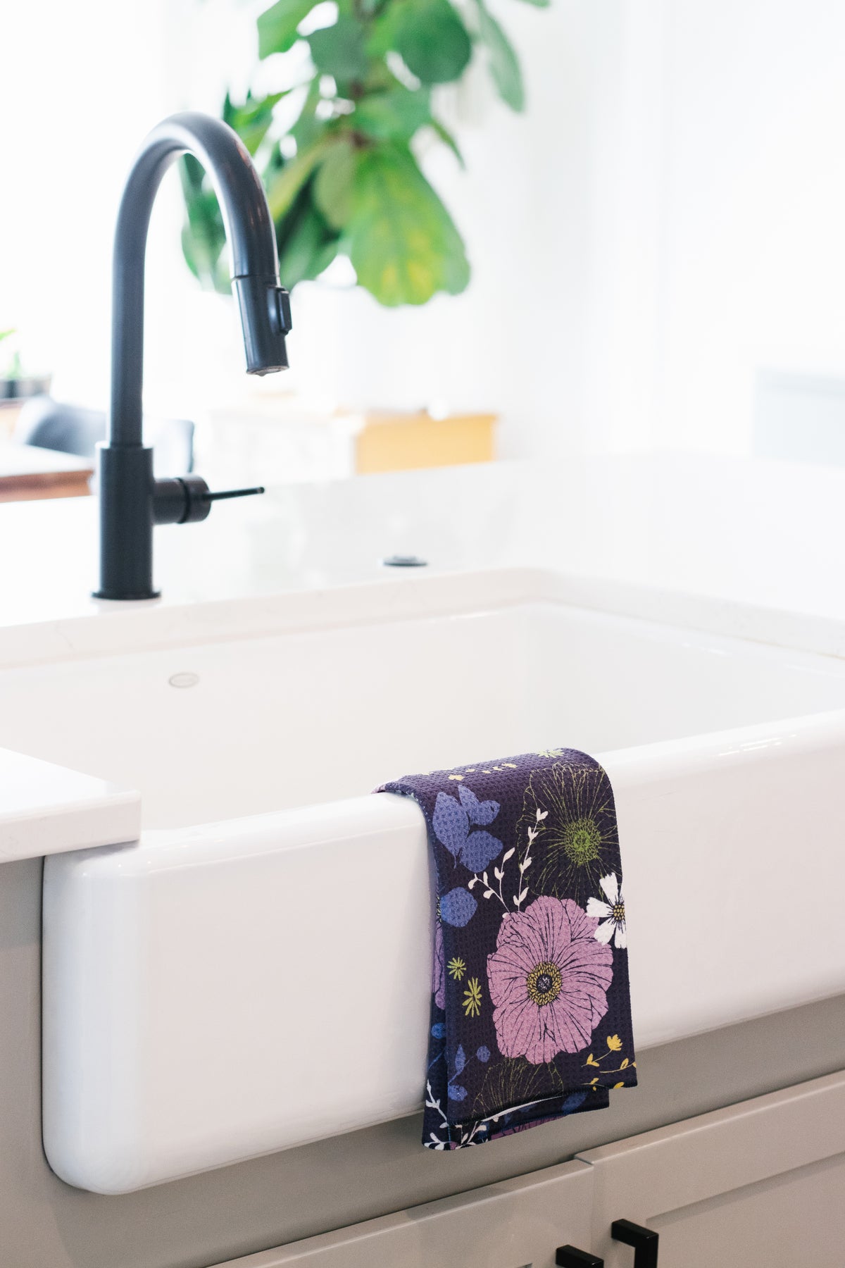 White farmhouse kitchen sink with a black faucet and a floral waffle-weave kitchen tea towel hanging on the side.