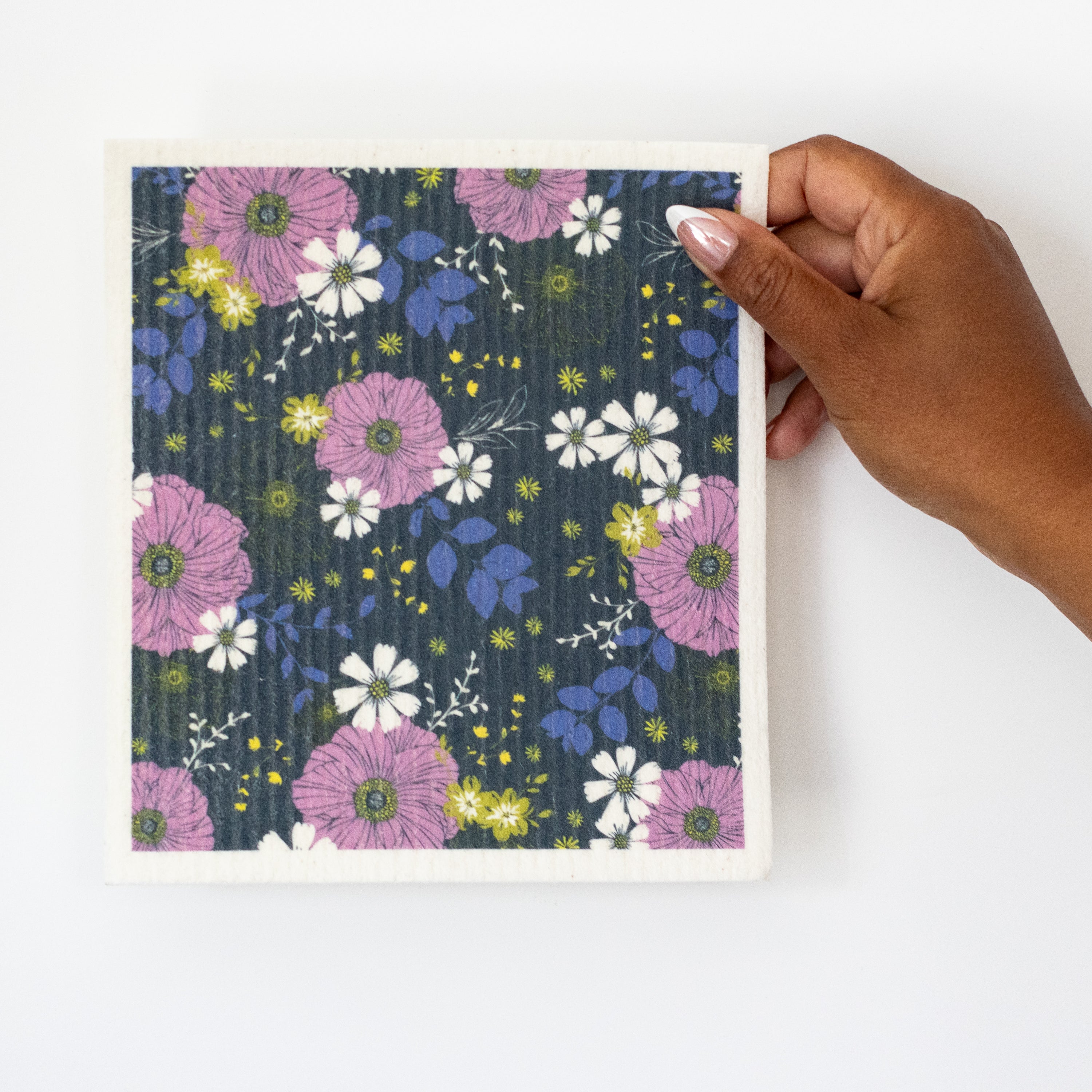 A hand holds a Swedish dishcloth featuring a floral pattern on a dark navy background with lavender purple, white, and lime green flowers and blue leaves, set against a plain white background.