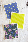 A Swedish dishcloth set of three including one with a hand drawn blue floral design, another with a floral design of purple, white, and green flowers with blue leaves on a dark background, and also a bright green and white striped reusable cleaning cloth laid flat on white background.