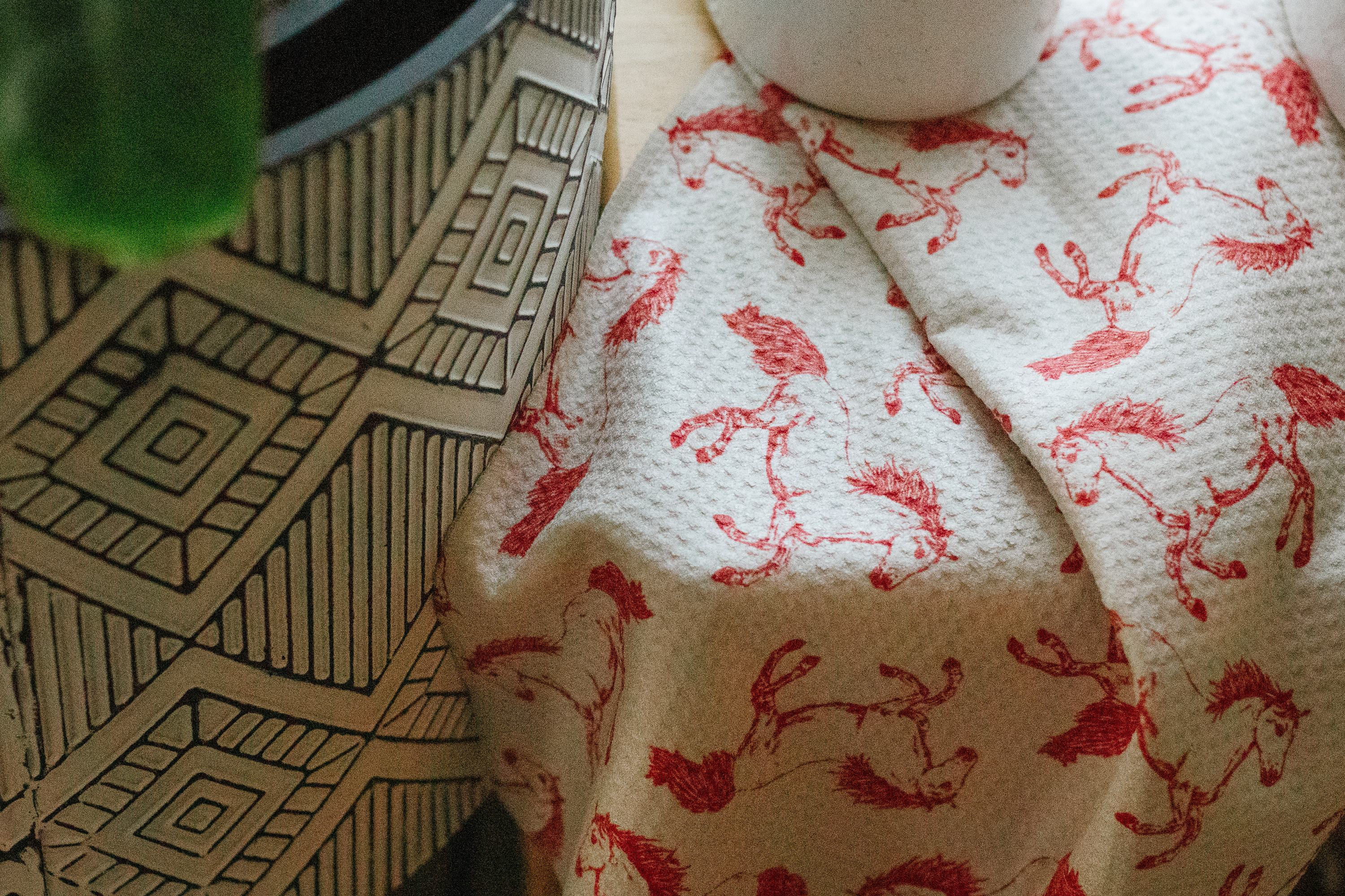 Decorative pot next to a waffle-weave kitchen tea towel with a red horse patternon a light beige background draped on a wood surface
