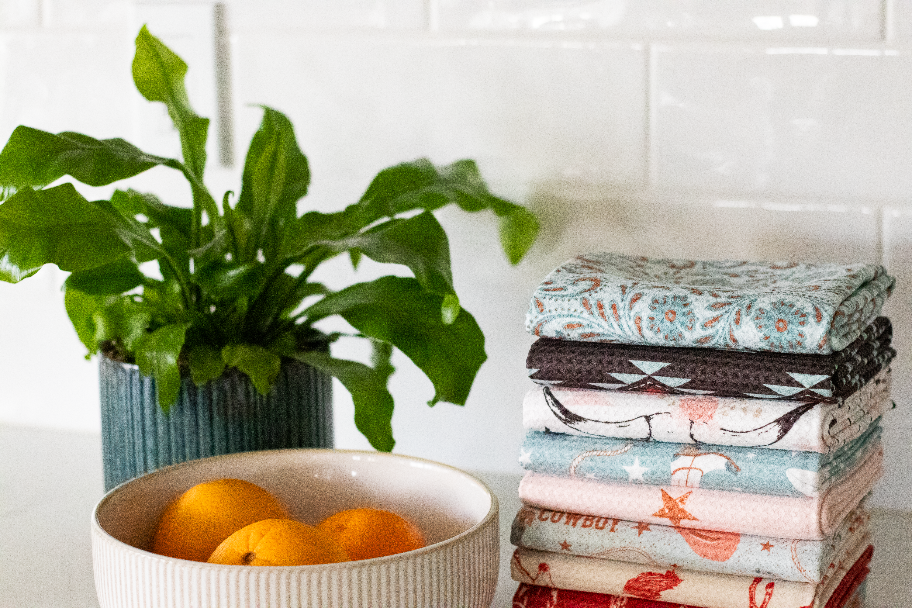 Stack of folded western-style waffle-weave kitchen tea towels with a bowl of oranges and a plant on a white surface and tile background