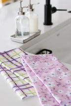 Two kitchen towels, one plaid and one floral, on a countertop by a sink with soap dispensers in the background.