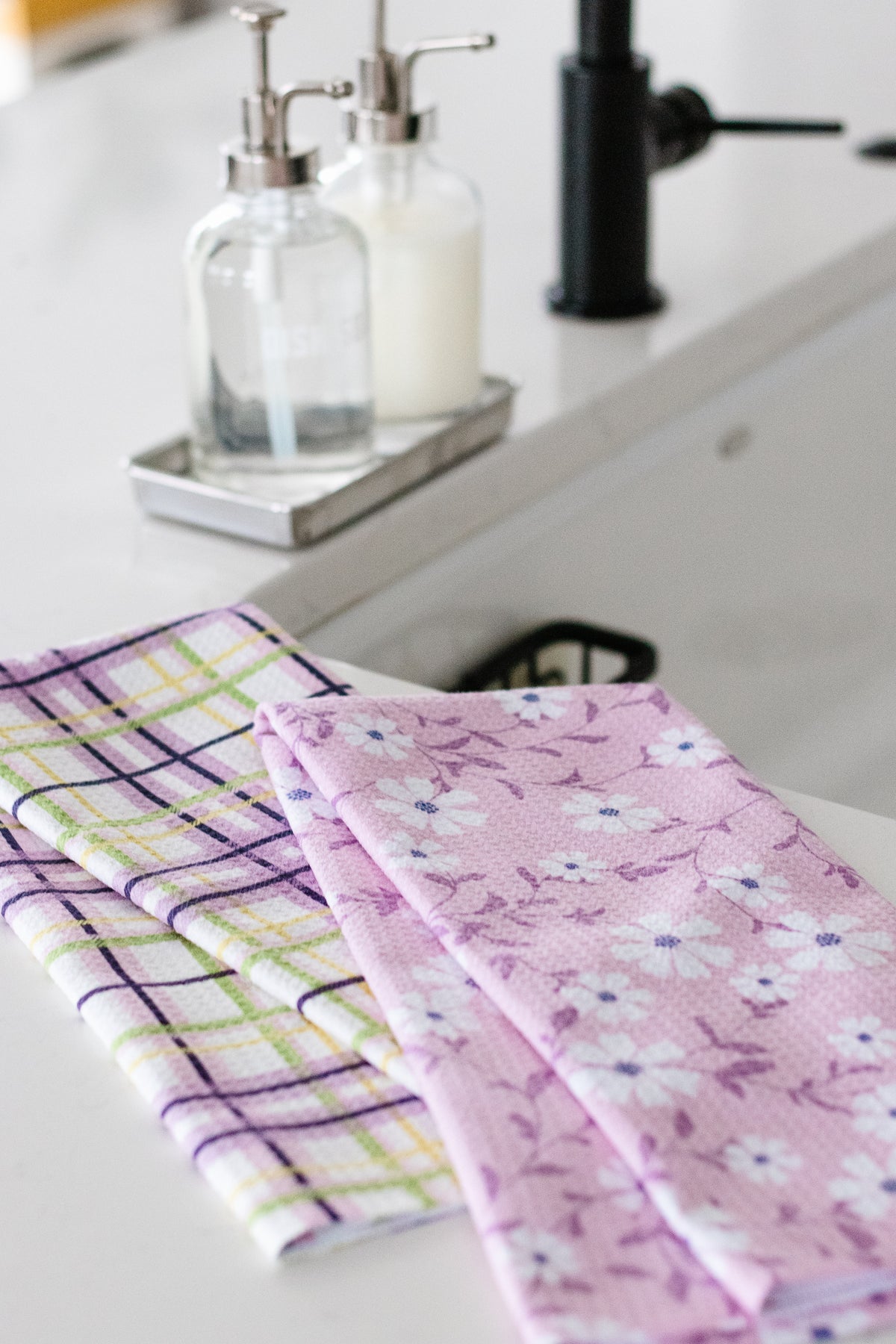 Two kitchen towels, one plaid and one floral, on a countertop by a sink with soap dispensers in the background.