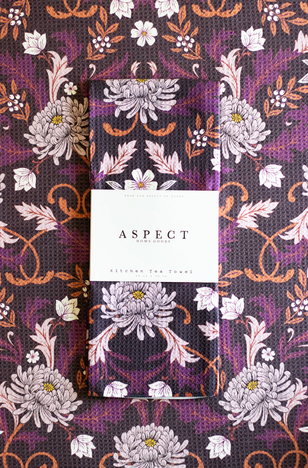 A folded waffle-weave kitchen tea towel with a purple, white, and orange floral pattern, featuring a white paper belly band label in the center that reads 'ASPECT HOME GOODS & Kitchen Tea Towel'.