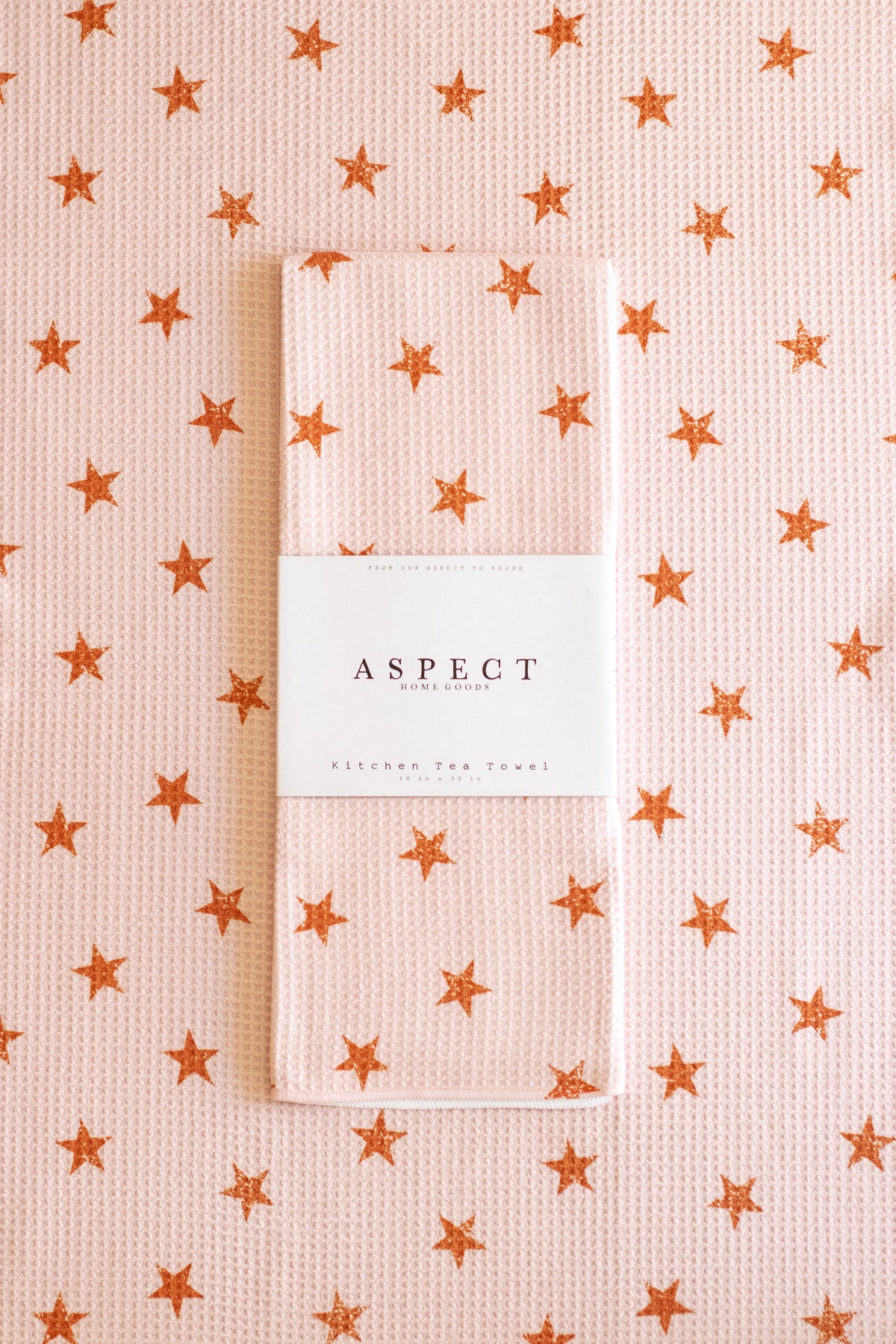 A folded waffle-weave kitchen tea towel with a light pink and brown distressed star pattern, wrapped in a white paper belly band labeled 'ASPECT HOME GOODS', set against a matching patterned background.