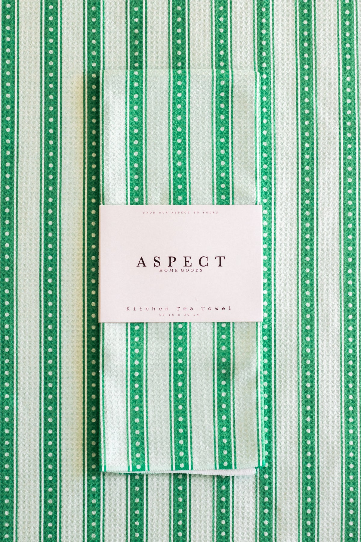 A folded kitchen tea towel with a green and white striped pattern, wrapped in a white paper belly band labeled 'ASPECT KITCHEN TEA TOWEL'. The background is a matching flat-lay of the same tea towel pattern.