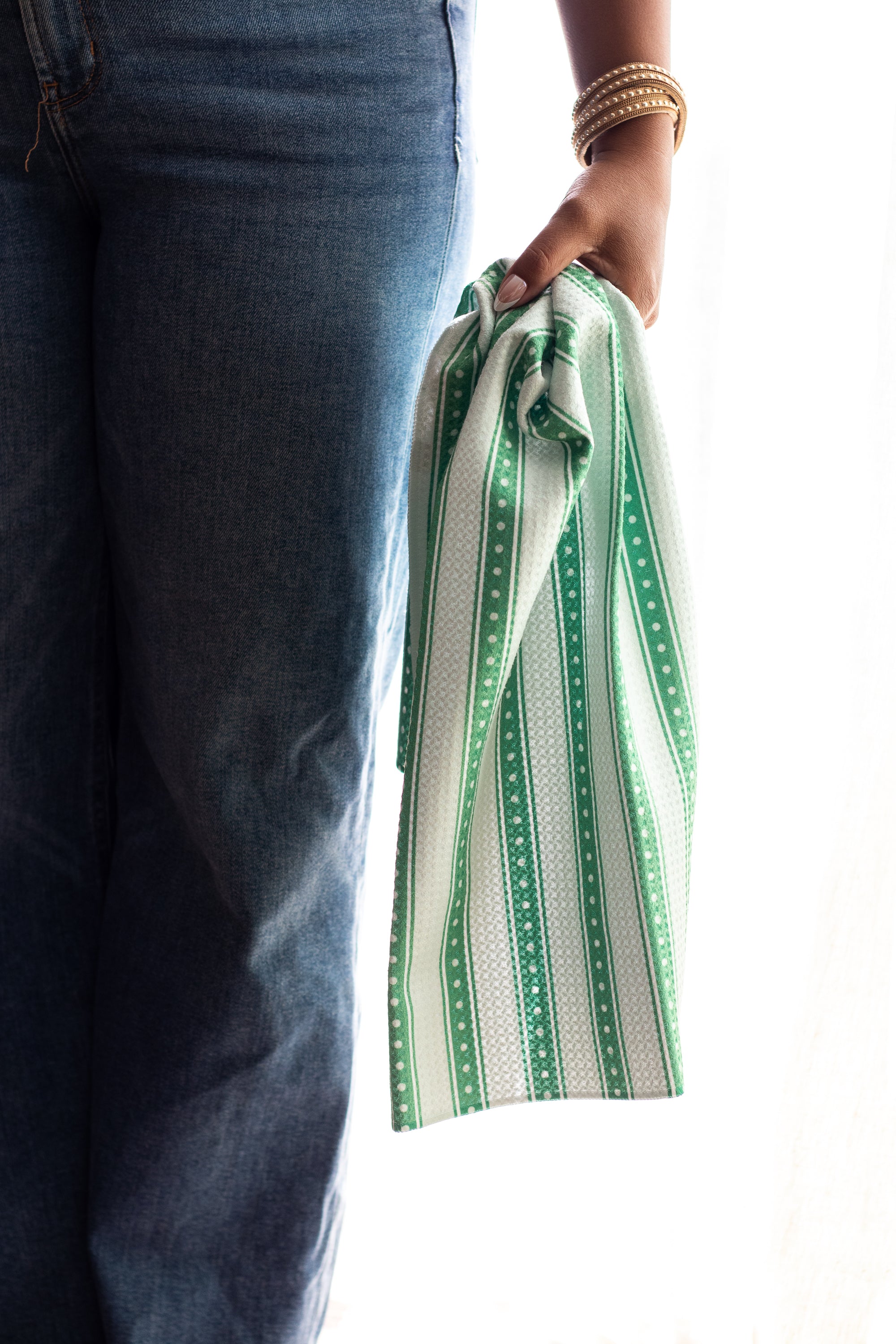 A person wearing blue jeans holds a green-and-white patterned kitchen towel against a bright white background.