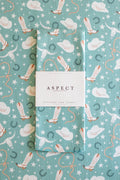 A folded waffle-weave kitchen towel with a western-themed print of cowgirl boots, lassos, cowgirl hats, horseshoes, and stars on a turquoise background, wrapped with a white paper belly band that reads 'ASPECT HOME GOODS' and 'KITCHEN TEA TOWEL', laid on a matching patterned background.