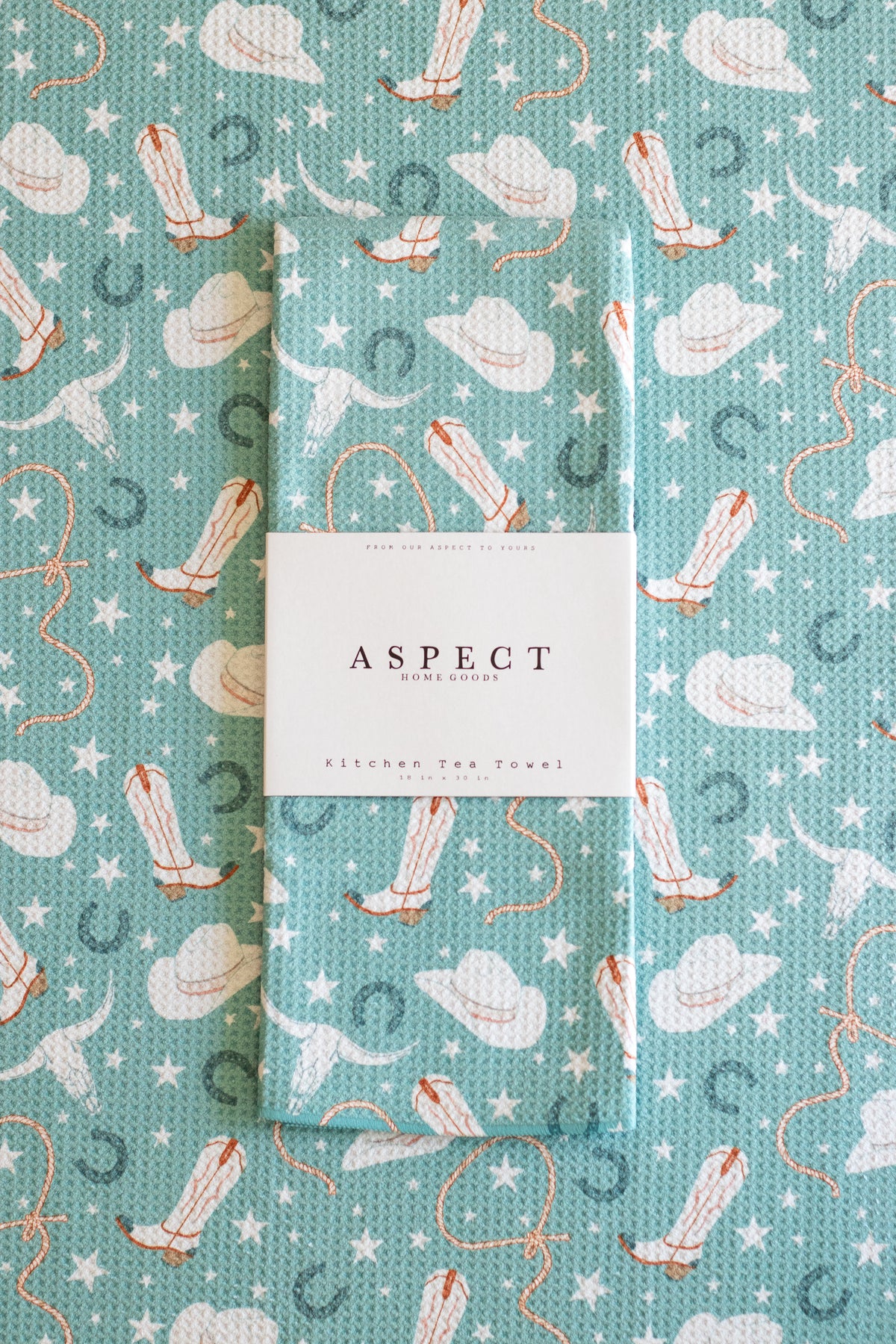 A folded waffle-weave kitchen towel with a western-themed print of cowgirl boots, lassos, cowgirl hats, horseshoes, and stars on a turquoise background, wrapped with a white paper belly band that reads 'ASPECT HOME GOODS' and 'KITCHEN TEA TOWEL', laid on a matching patterned background.