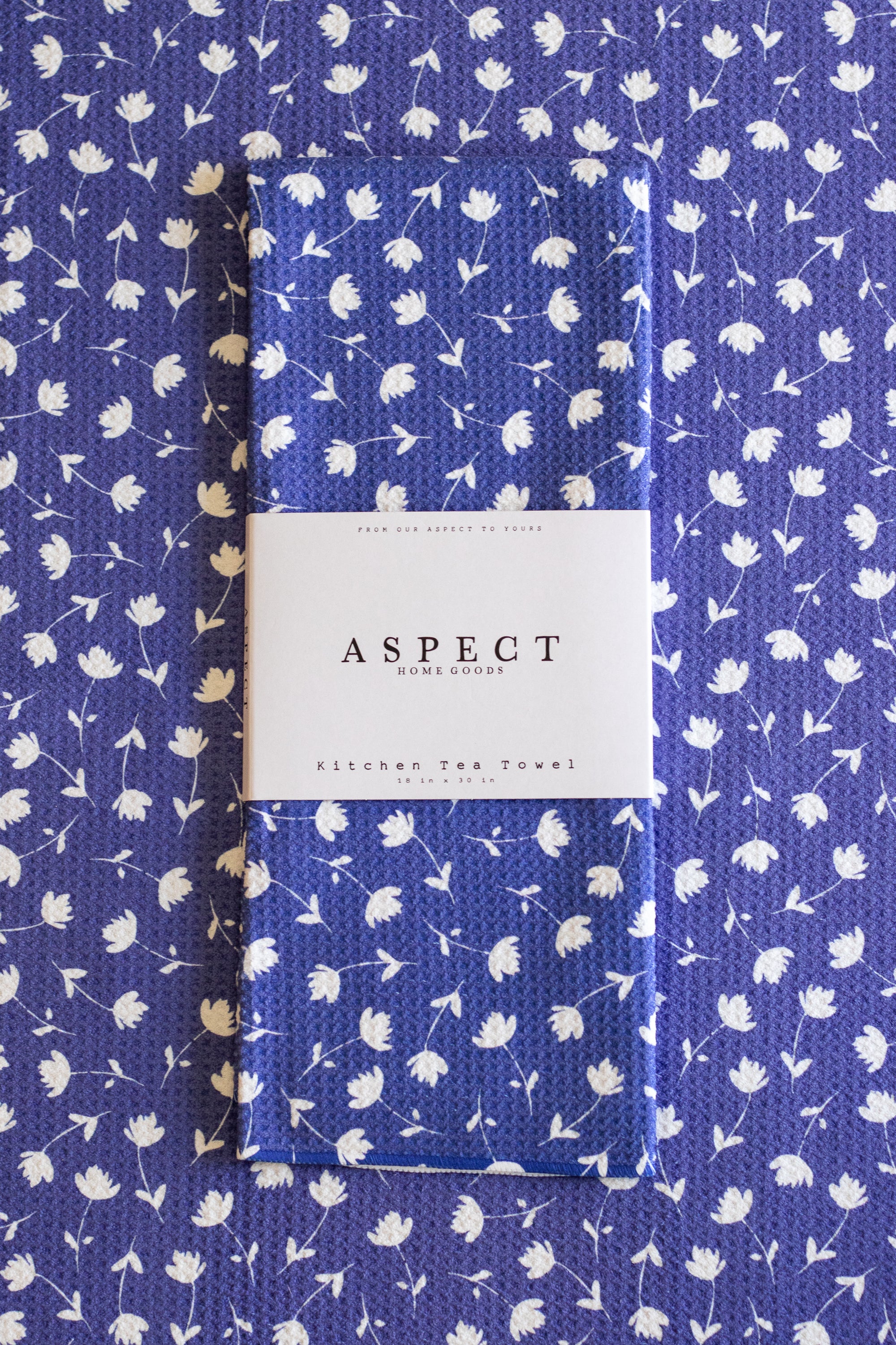 A folded waffle-weave kitchen tea towel with an indigo blue and white floral pattern, wrapped in a white paper belly band labeled 'ASPECT HOME GOODS' and 'kitchen tea towel', laid flat on a matching patterned blue surface.