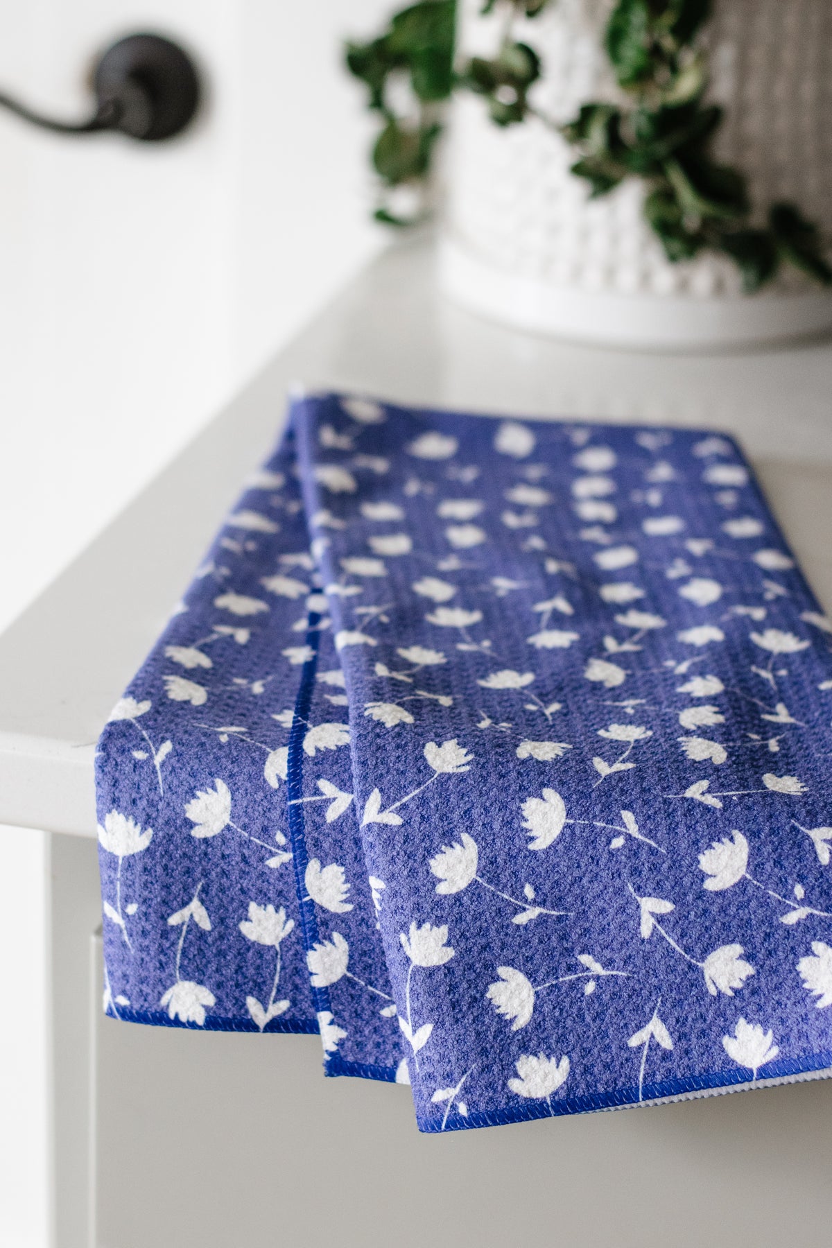 Blue waffle-weave kitchen tea towel with a white floral pattern on a white surface and a plant in the background.