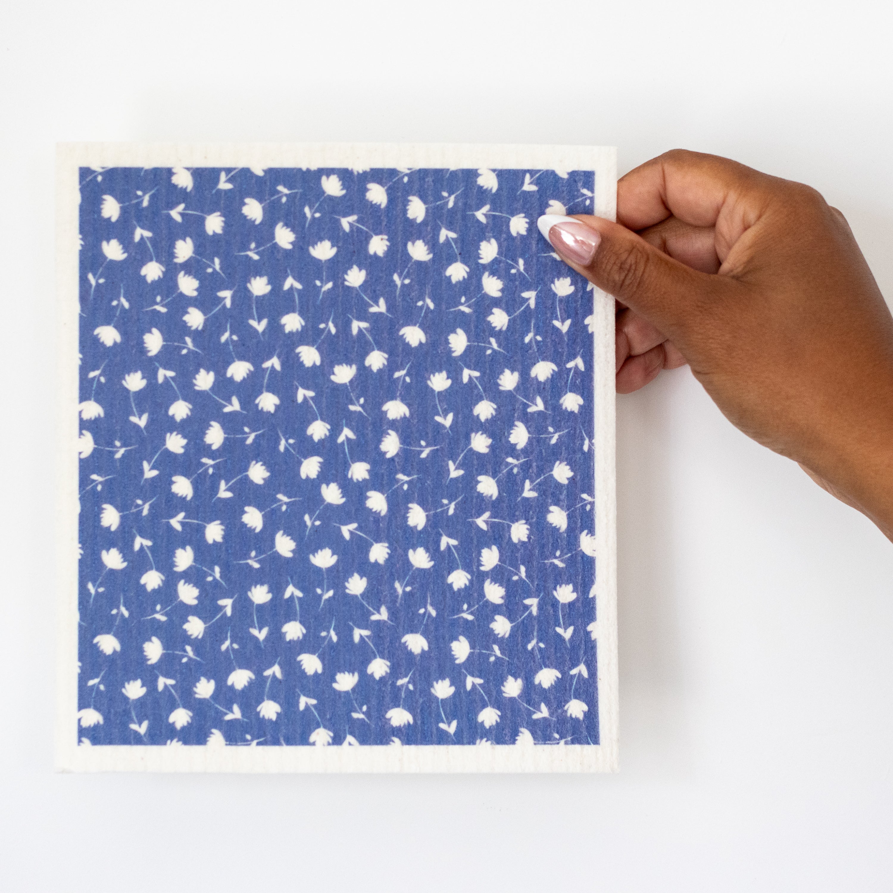 A hand holding a Swedish dishcloth with white hand-painted flower pattern on a blue background.