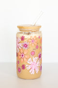 A can glass with a bamboo lid and glass straw containing a coffee drink is decorated with a colorful floral design in pink, orange, and purple against a white background.