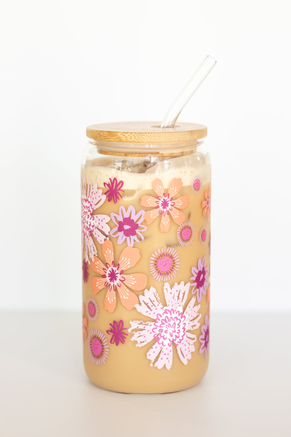 A can glass with a bamboo lid and glass straw containing a coffee drink is decorated with a colorful floral design in pink, orange, and purple against a white background.