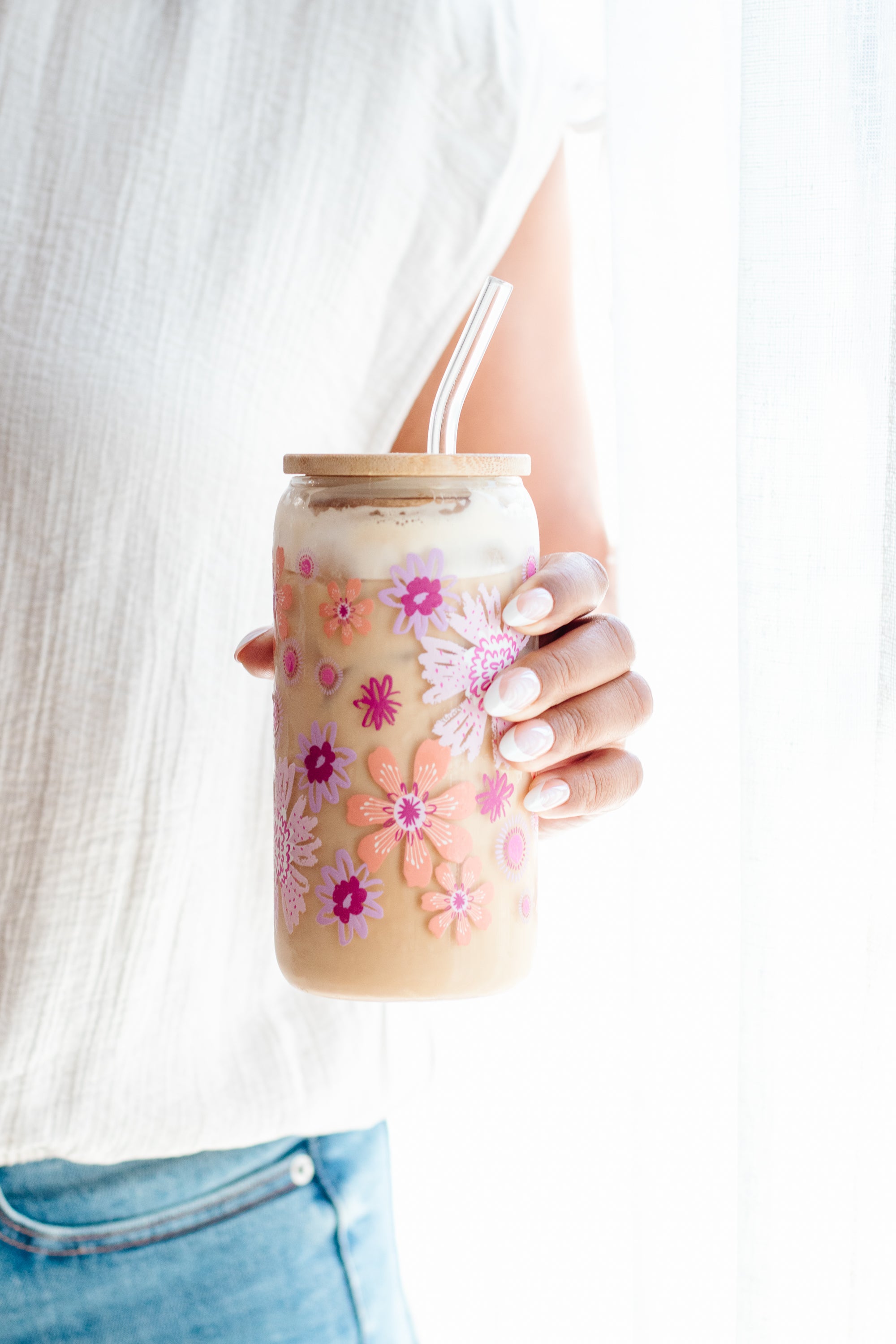 A person in a white top and blue jeans holds a clear can glass tumbler with a light wood lid and two straws. The tumbler is decorated with pink, orange, and purple floral patterns and contains an iced coffee drink.