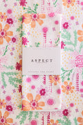 A folded waffle-weave kitchen tea towel with a colorful floral and botanical print, wrapped in a white paper belly band labeled 'ASPECT HOME GOODS'. The background is a matching flat-lay of the same tea towel pattern.