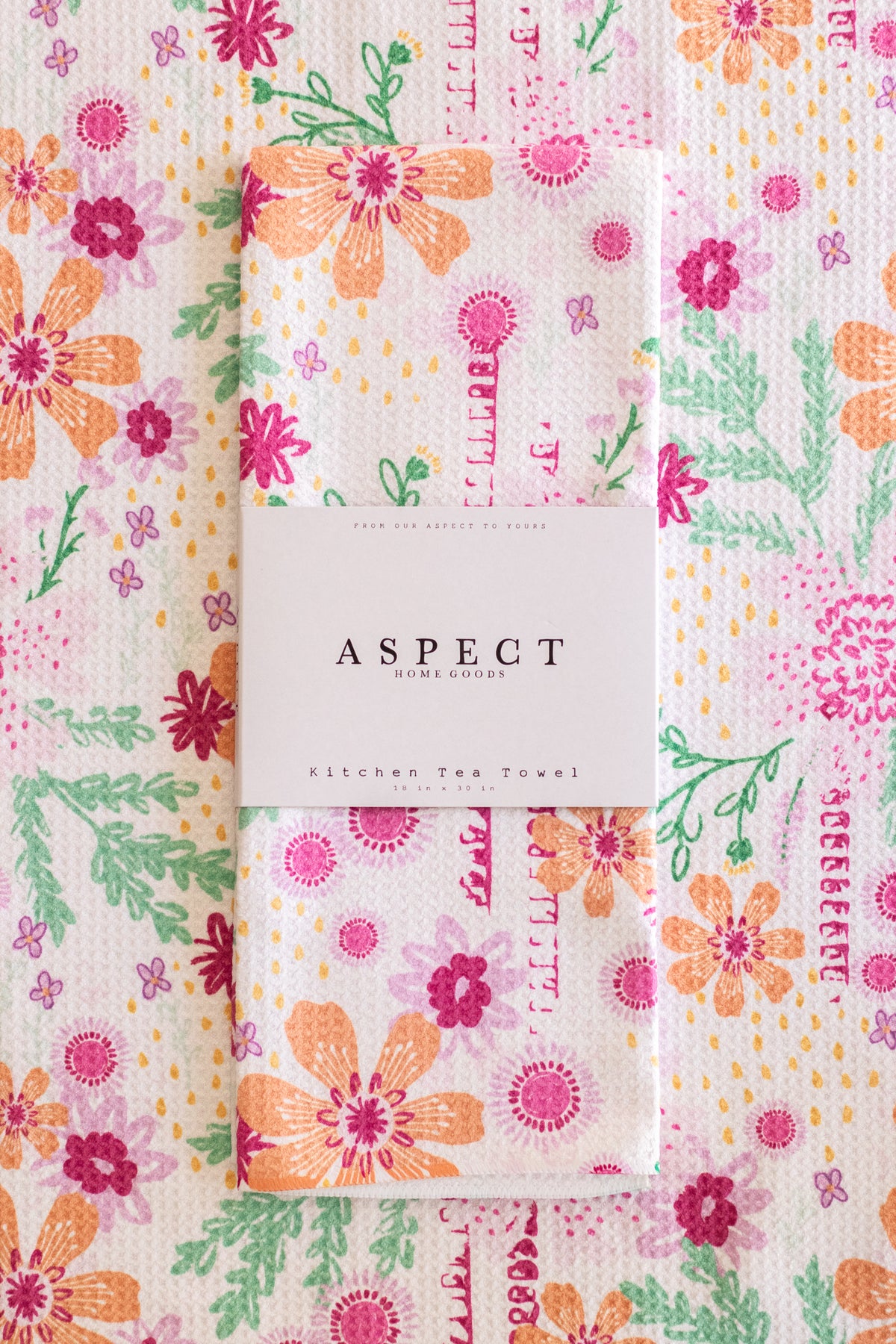 A folded waffle-weave kitchen tea towel with a colorful floral and botanical print, wrapped in a white paper belly band labeled 'ASPECT HOME GOODS'. The background is a matching flat-lay of the same tea towel pattern.