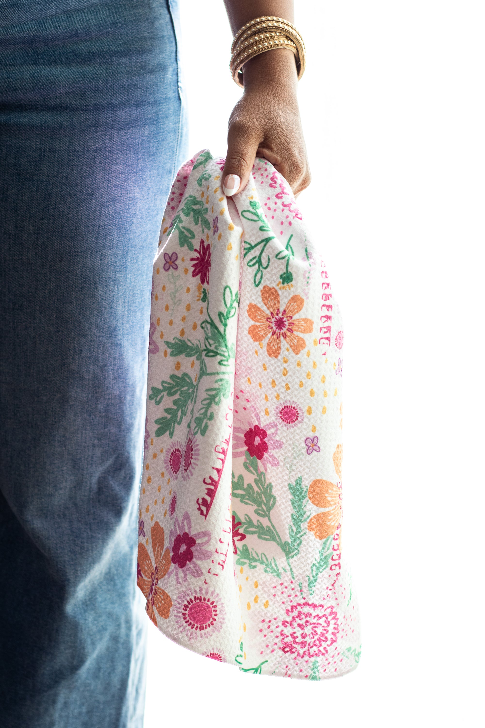 A person wearing blue jeans holds a waffle-weave kitchen tea towel with a colorful floral-and-botanical print on a white background. The print includes pink, orange, and purple flowers with greenery and yellow accents.