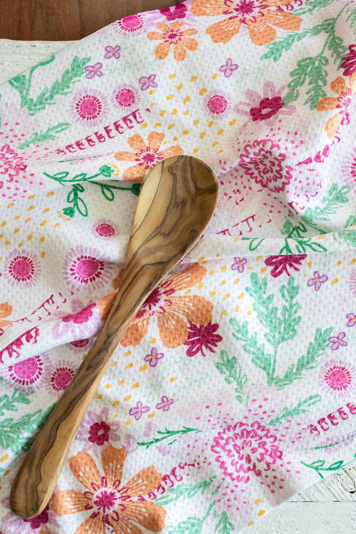 A wooden spoon with a natural olive wood grain pattern rests on a white tea towel printed with colorful pink, orange, and green floral motifs.