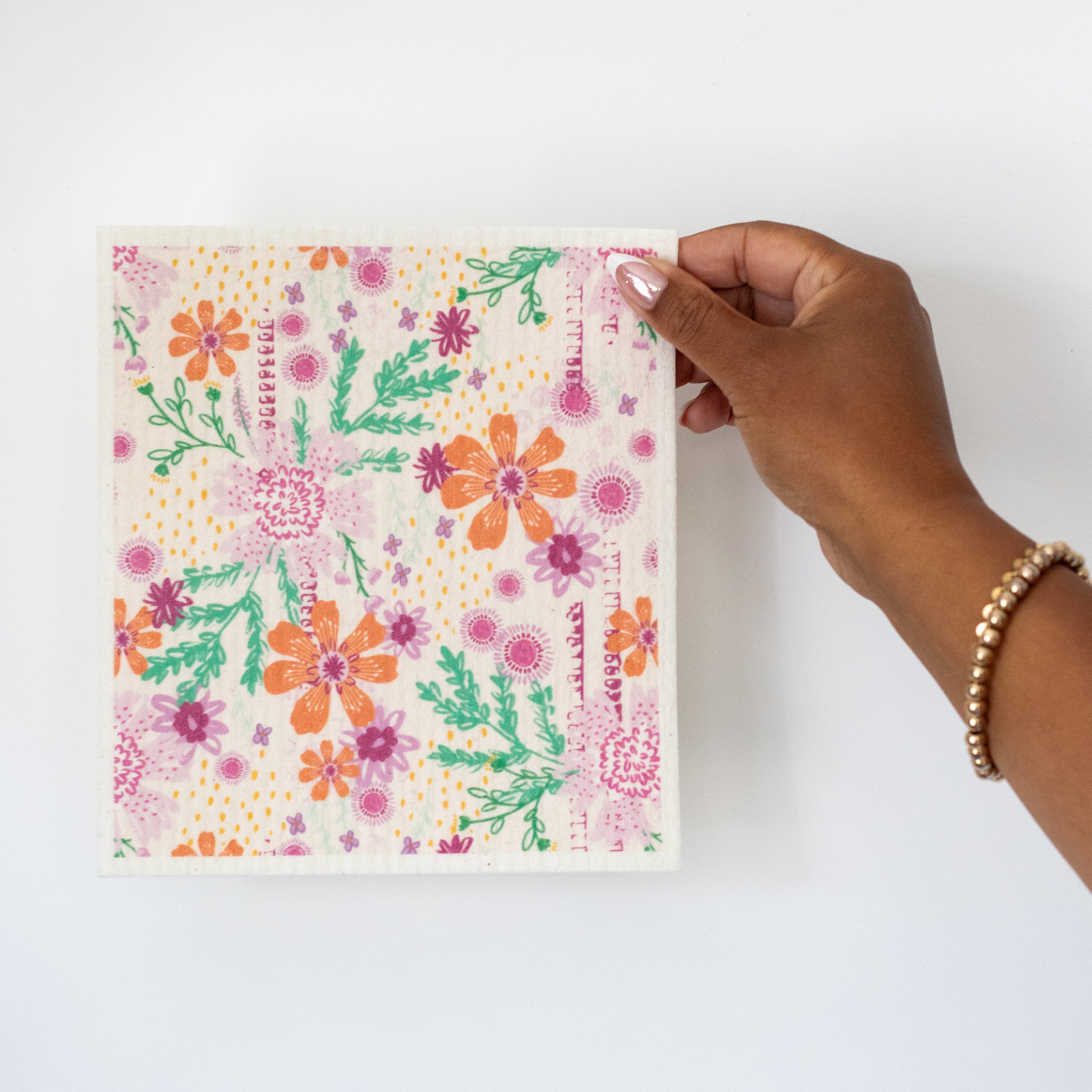 A hand holding a Swedish dishcloth featuring a colorful floral print with orange, pink, and purple flowers and greenery against a white background.