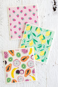 Three Swedish dishcloths featuring colorful fruit and vegetable patterns (strawberries, lemons, and assorted fruits) on a white textured wooden background.