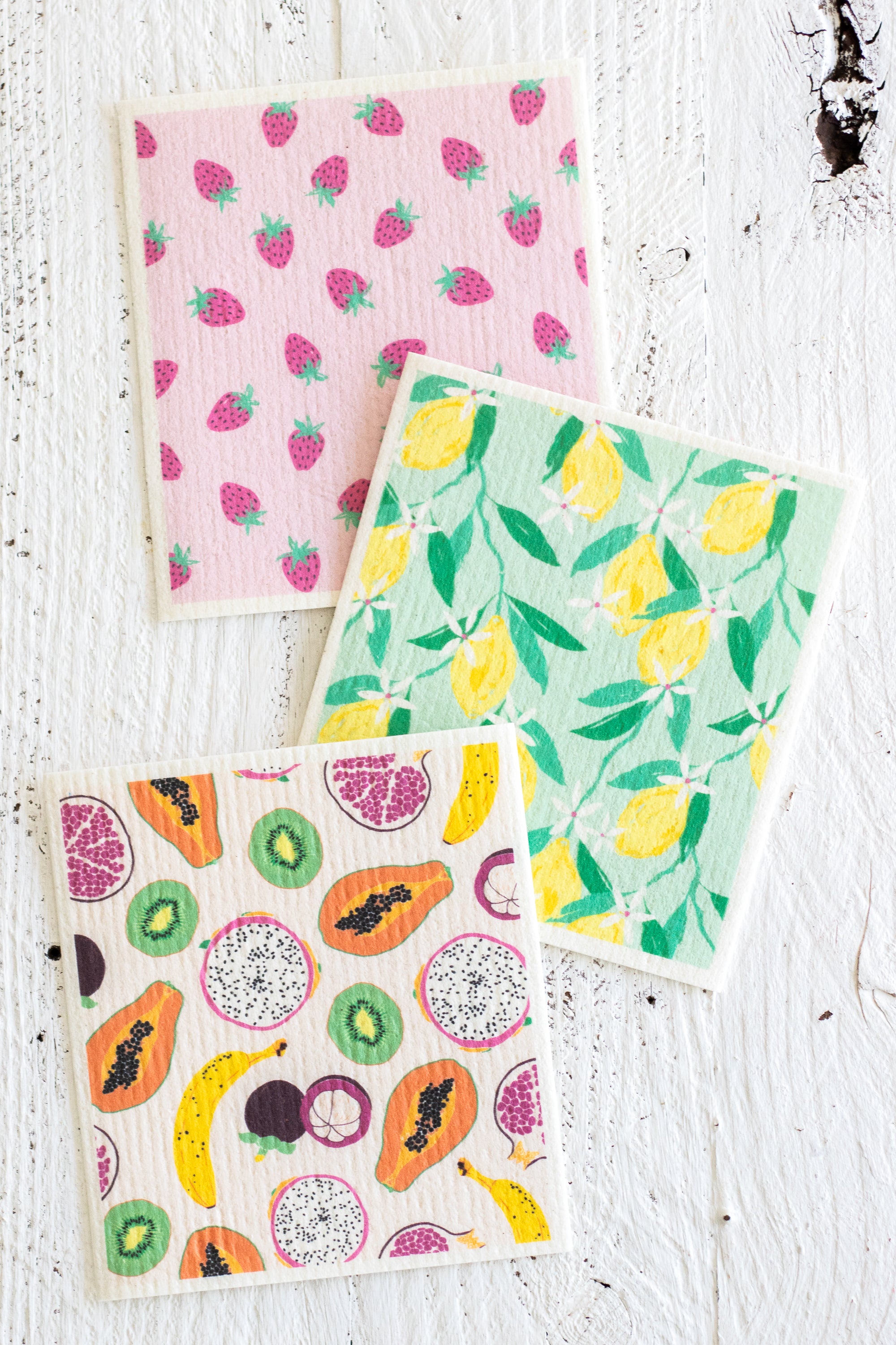 Three Swedish dishcloths featuring colorful fruit and vegetable patterns (strawberries, lemons, and assorted fruits) on a white textured wooden background.
