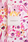 A folded kitchen tea towel with a colorful floral print is wrapped in a white paper belly band labeled 'ASPECT HOME GOODS KITCHEN TEA TOWEL'. The towel features a vibrant pattern of pink, purple, orange, and yellow flowers on a white background, laid flat on a matching patterned surface.