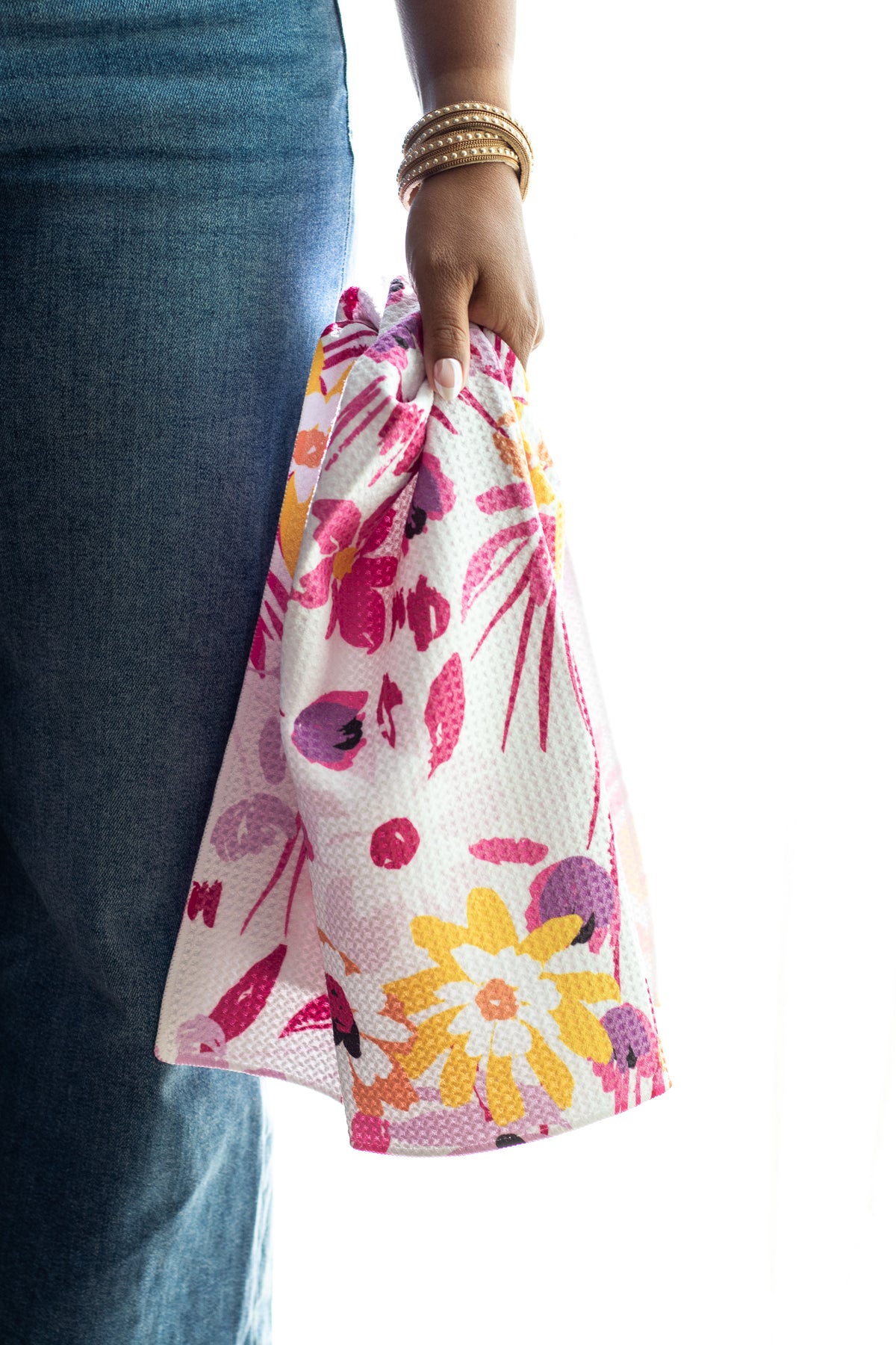 A person wearing blue jeans holds a waffle-weave kitchen tea towel with a colorful floral print in pink, yellow, and orange against a white background.
