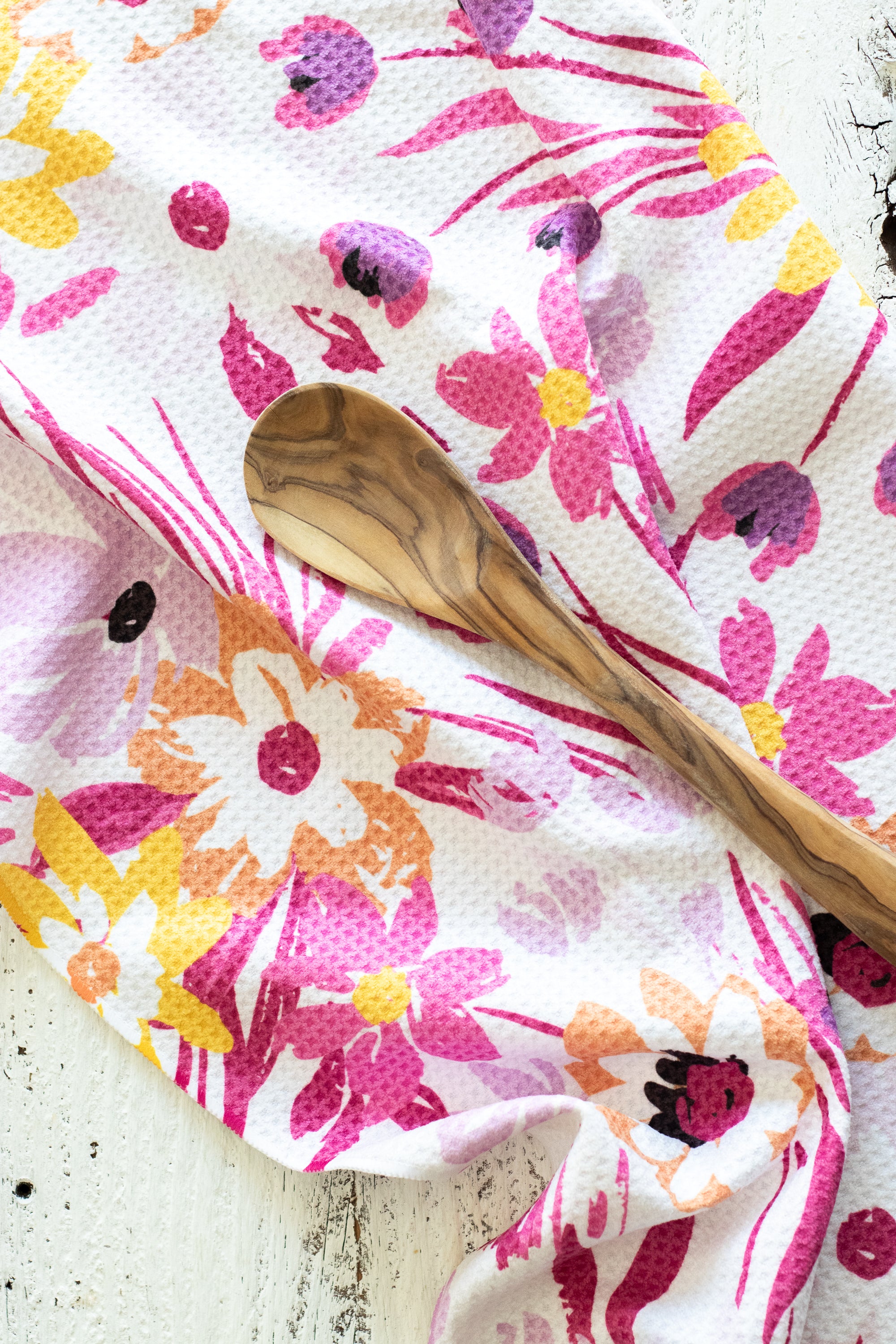 A close-up of a waffle-weave kitchen tea towel or cloth with a colorful floral print in pink, purple, orange, and yellow on a white background, draped over a light wood spoon on a white rustic surface.