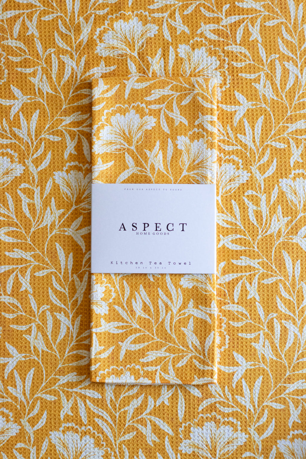 A folded waffle-weave kitchen tea towel with a white botanical leaf pattern on a mustard yellow background, wrapped in a white paper belly band labeled 'ASPECT HOME GOODS' and laid on a towel with the same pattern.