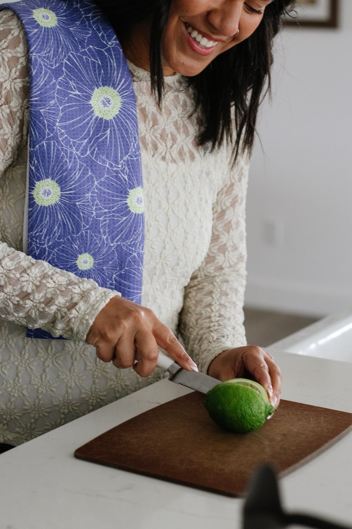 Woman cutting a lime on a wooden cutting board with a blue-and-lime-green floral-patterned waffle-weave kitchen tea towel draped over her shoulder.