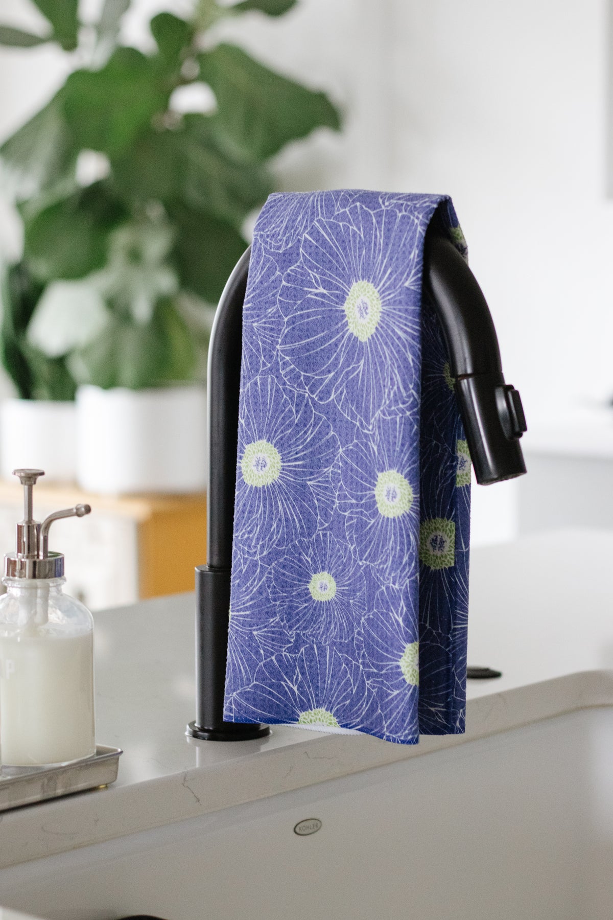 Blue-and-lime-green floral-patterned waffle-weave kitchen tea towel hanging on a black faucet over a sink next to a soap dispenser, with a plant in the background.