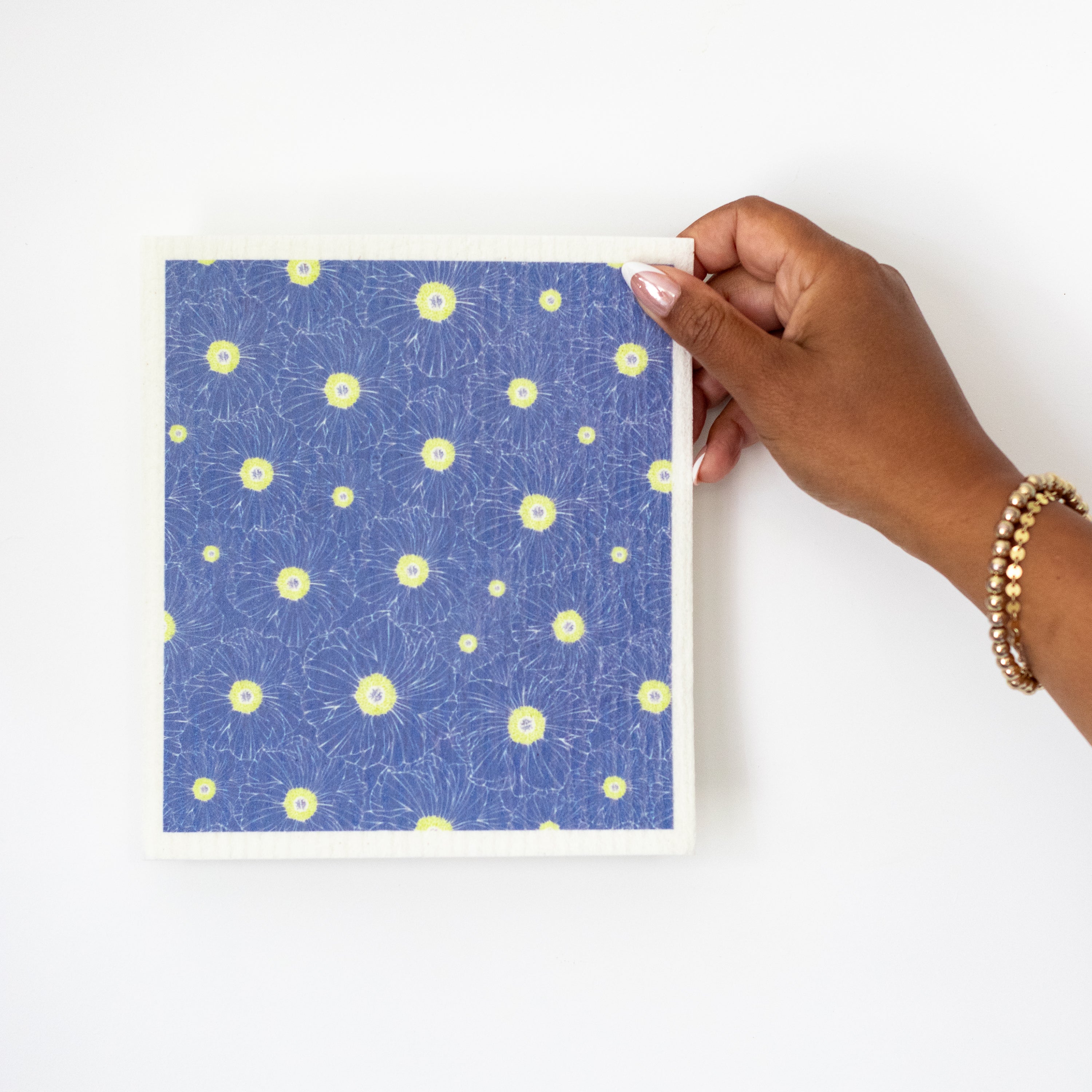 A hand holding a Swedish dishcloth with a hand drawn Anemone flower pattern in bright green on a blue background.