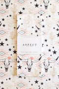 A folded waffle-weave kitchen tea towel from Aspect, featuring a southwestern pattern with black stars, beige cacti, and black-and-white longhorn cow skulls with pink geometric and floral accents on a cream background. The towel is wrapped in a white paper sleeve with the brand name 'ASPECT HOME GOODS' printed on it, and is laid flat on top of a matching patterned fabric.