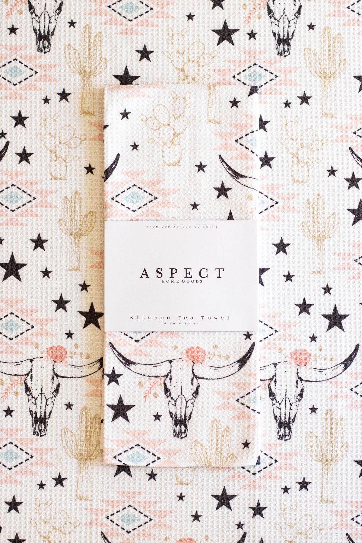A folded waffle-weave kitchen tea towel from Aspect, featuring a southwestern pattern with black stars, beige cacti, and black-and-white longhorn cow skulls with pink geometric and floral accents on a cream background. The towel is wrapped in a white paper sleeve with the brand name 'ASPECT HOME GOODS' printed on it, and is laid flat on top of a matching patterned fabric.