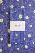 A folded waffle-weave kitchen tea towel with an indigo blue, lime green, and white hand-drawn floral pattern, wrapped in a white paper belly band labeled 'ASPECT HOME GOODS', placed on top of a matching patterned background.
