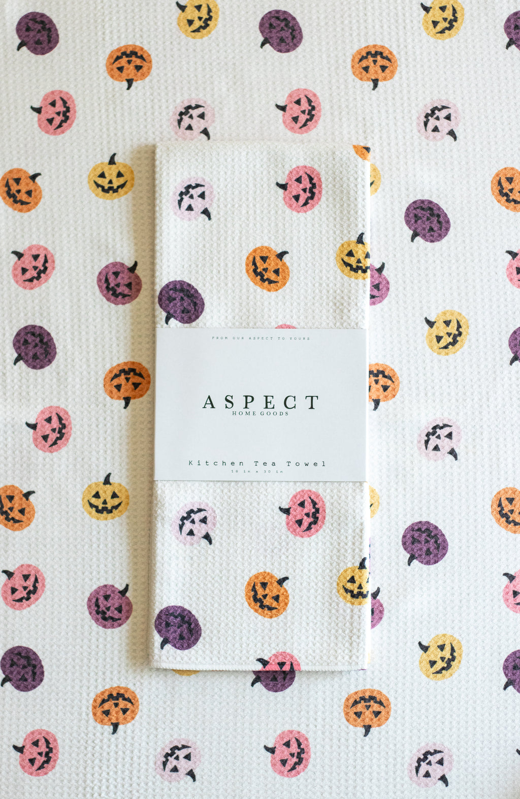 A folded white waffle-weave kitchen tea towel with a repeating pattern of jack-'o'-lantern pumpkins in orange, pink, purple and yellow, wrapped in a paper belly band labeled 'ASPECT HOME GOODS & KITCHEN TEA TOWEL', on top of another towel with the same pattern.