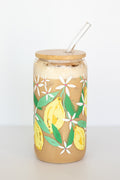 A can glass that is decorated with a watercolor lemon design, with a wooden lid and a glass straw, containing a coffee drink, against a white background.