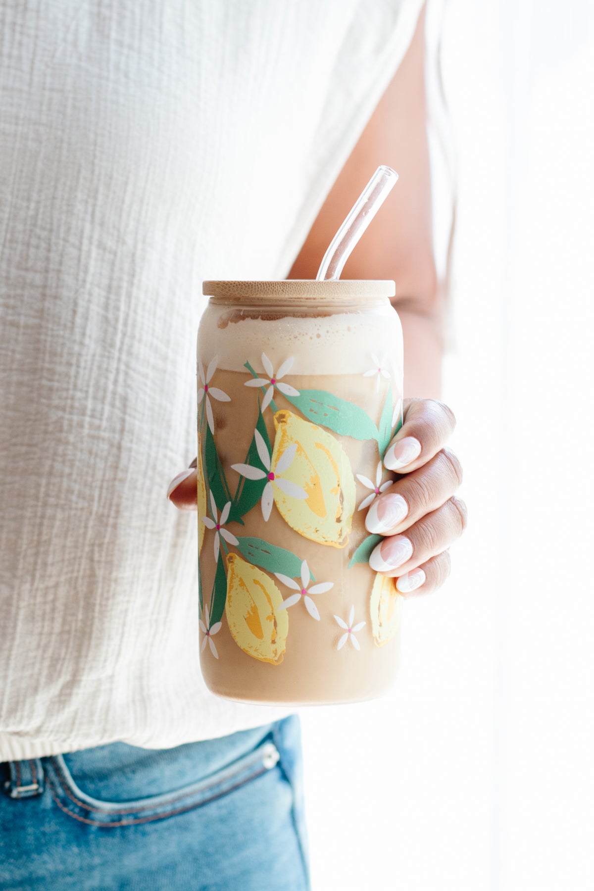 A person holding a clear glass can-style tumbler with a bamboo lid and glass straw, filled with an iced coffee drink. The tumbler is decorated with watercolor yellow lemons and white flowers with green leaves.