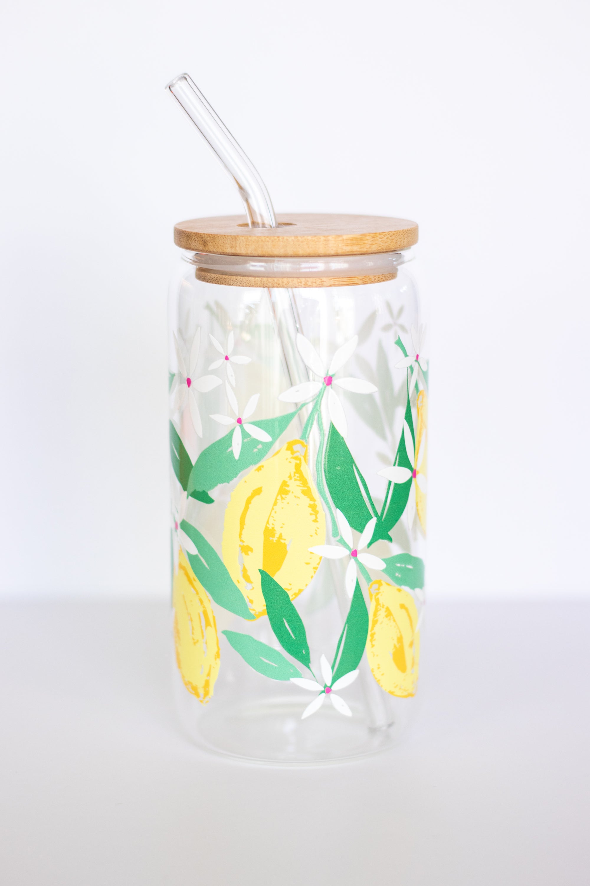 A can glass that is decorated with a watercolor lemon design, with a wooden lid and a glass straw, against a white background.