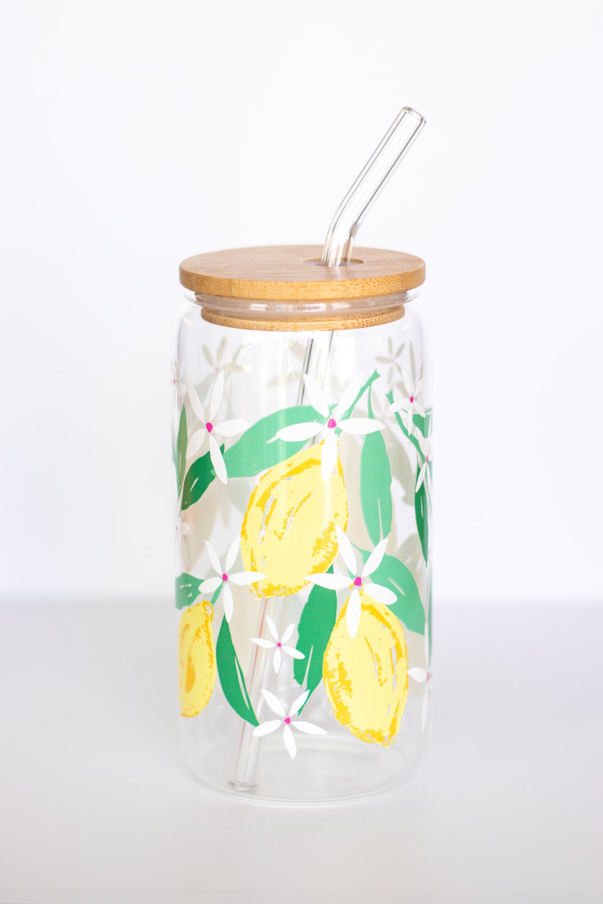 A can glass that is decorated with a watercolor lemon design, with a wooden lid and a glass straw, against a white background.