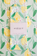 A folded waffle-weave kitchen tea towel with a lemon-and-leaf pattern, wrapped in a white paper belly band labeled 'ASPECT HOME GOODS' and 'kitchen tea towel', laid flat on a matching patterned background.